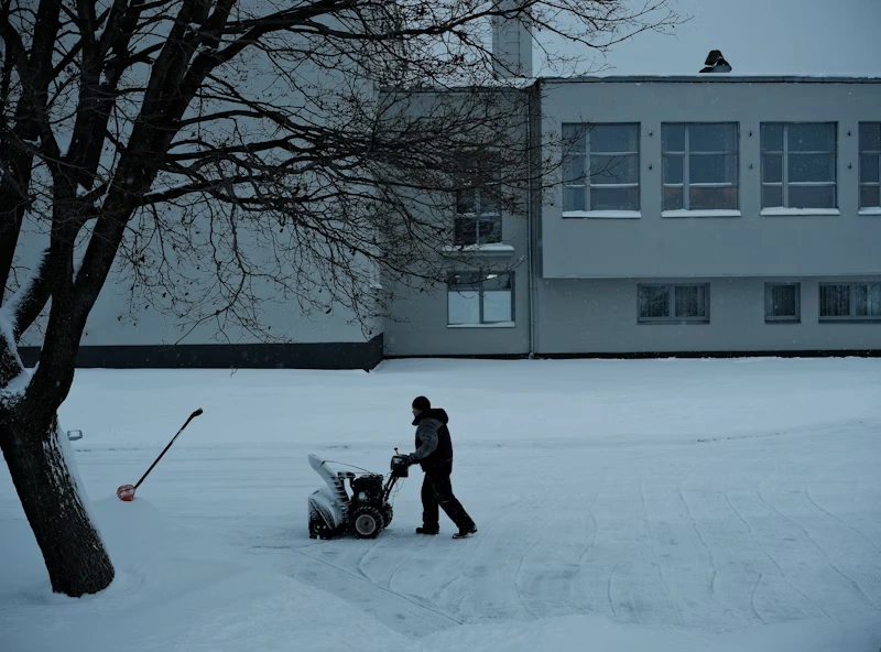 Person operating a snow blower on a snow-covered sidewalk in front of a large building during winter. A snow shovel leans against a nearby tree, and bare branches hang overhead.