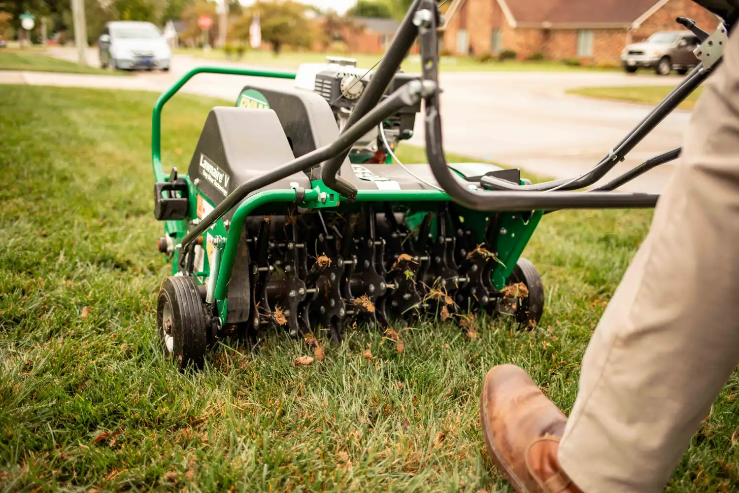 Close-up of a lawn care professional using a green stand-on core aerator machine on a residential lawn. Soil plugs are being pulled from the grass as the machine operates near a neighborhood street.