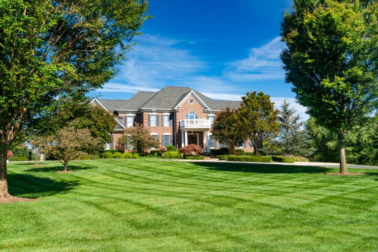 Large brick house with a well-manicured lawn featuring diagonal mowing stripes. Mature trees and neatly trimmed shrubs frame the yard under a clear blue sky.