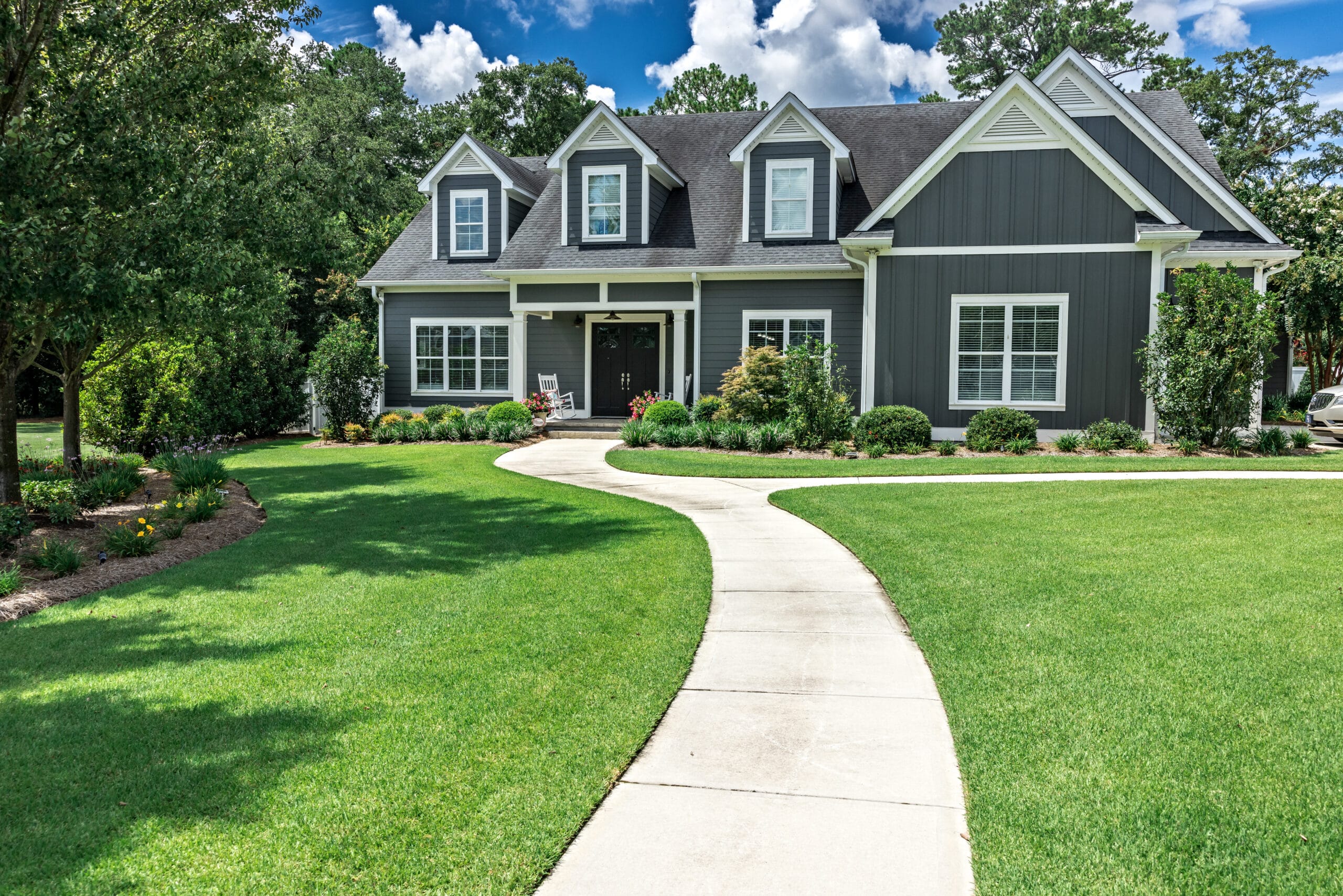 A beautiful gray suburban home with white trim and dormer windows, set behind a curved concrete walkway leading through a lush, green lawn. Well-maintained landscaping with neatly trimmed bushes and flower beds borders the house, and mature trees frame the yard under a bright blue sky with scattered clouds.