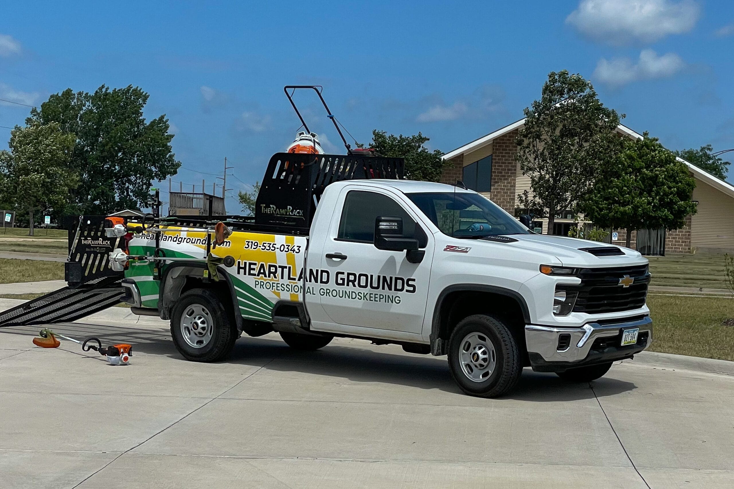 Heartland Grounds work truck parked on a paved surface, equipped with a Ramp Rack and lawn care equipment including a mower and trimmer. The truck features company branding, contact info, and a website URL.