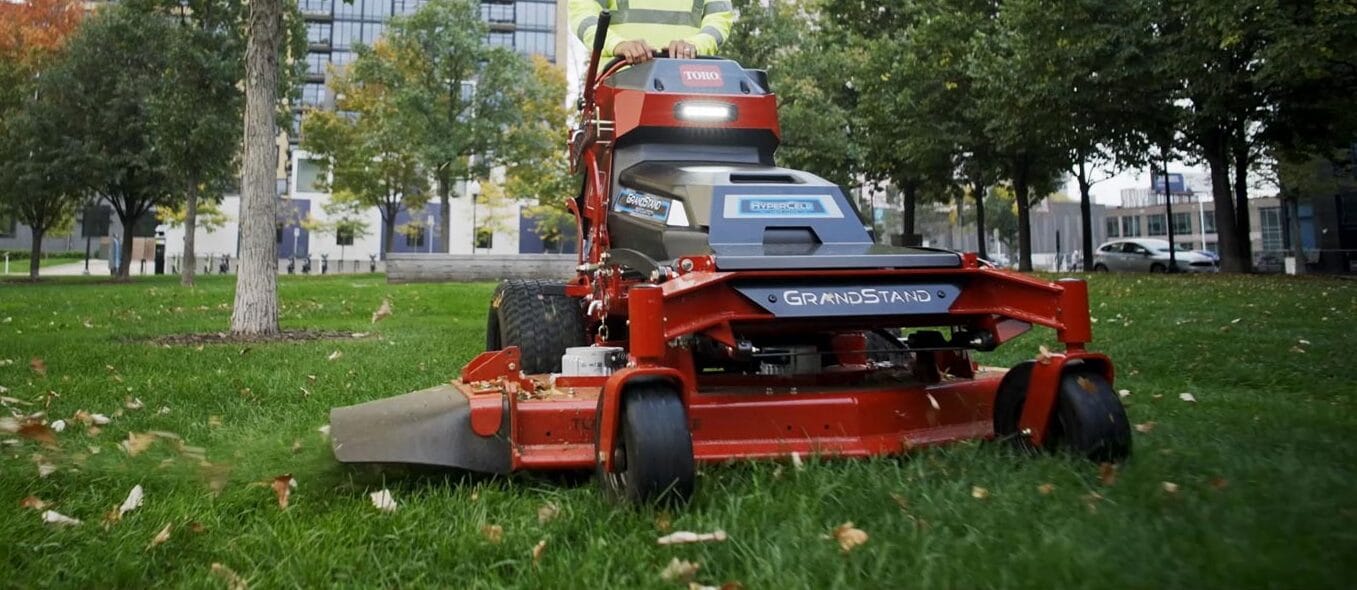 Commercial stand-on lawn mower cutting grass in a park near buildings and trees in Iowa City.
