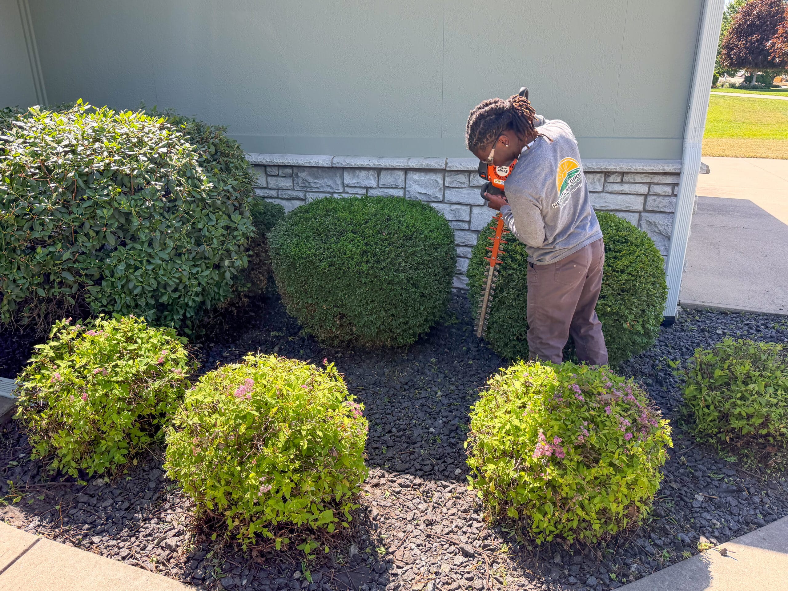 Heartland Grounds employee trimming shrubs in a residential landscape bed with decorative rock mulch beside a home, shaping neatly maintained green bushes.